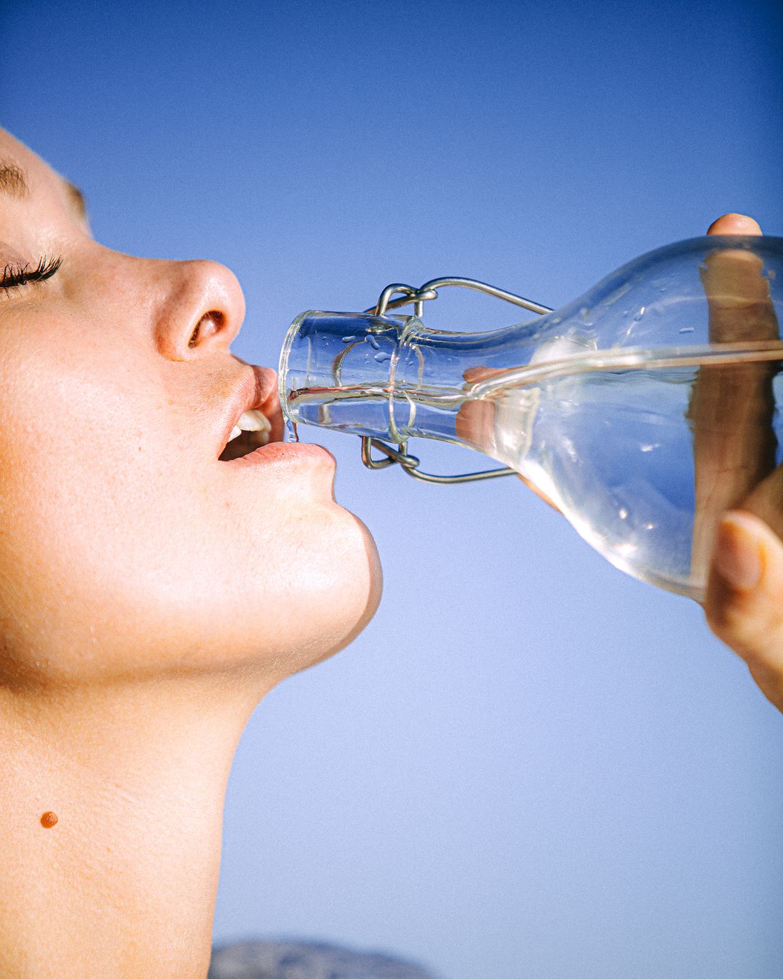 Woman drinking water from a glass bottle against a clear sky